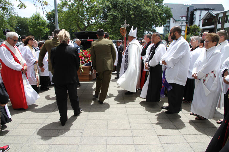 Whānau and clergy stand to honour Bishop Richard as Defence Force personnel carry him for his return to Wairewa.