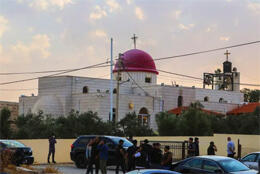 On a desert evening police and media staff stand outside a stone church with dome and bell tower 
