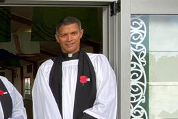 A Māori priest in choir dress with an Anzac poppy stands in a doorway flanked with a vertical kowhaiwhai panel