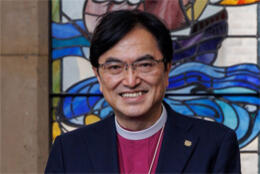A Japanese man with glasses smiles in front of a stained glass window depicting a sailing ship
