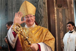 A middle aged male bishop in gold vestments waves at crowds