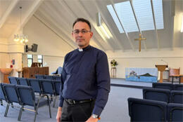 A male vicar in clericals stands in a 20th century church interior