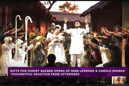 A stage full of Ghanaians in traditional dress includes a bearded figure in white and a woman in white holding a baby