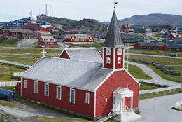 A 19th century white and reddish wooden Danish style church sits on grass with houses and rocky mountains behind