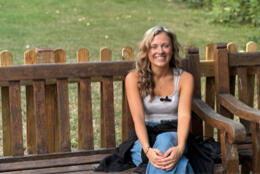 A young woman with blond curly hair smiles seated on a park bench