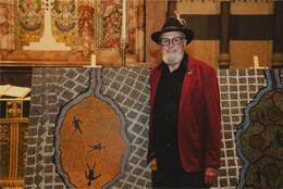 A senior man with white beard stands before two large Aboriginal dot landscape paintings inside an ornate stone church