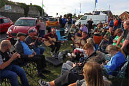 A circle of Irish musicians play fiddles, whistles and drums as the sun shines on a Northern town
