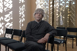 Anglican priest with curly grey hair sits in church lit with dappled light from modern patterned windows