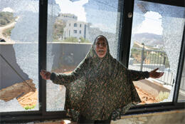 A Palestinian women in hijab gestures in horror before three large smashed windows in an upstairs apartment