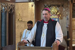 Jerusalem's Palestinian Archbishop stands in elevated wooden pulpit in black preaching scarf and rochet