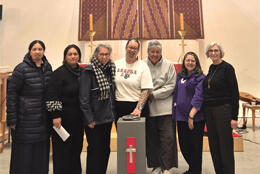 Bishop Susan Wallace stands in Christchurch Transitional Cathedral before a tukutuku panel, hand on a pounamu stone flanked by six female Anglican leaders