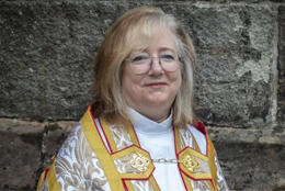 A blond haired female bishop in white robes and a gold cope smiles into the camera