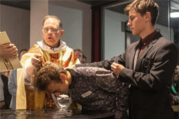 A young man is baptised by a Catholic priest supported by a friend 