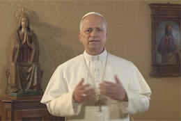 Pope Leo stand in white cassock against dark background and a statue of Mary