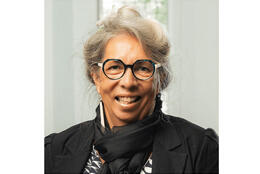 A portrait of Dr Jenny Te Paa smiling with pounamu framed glasses, bone earrings and hair in a bun.