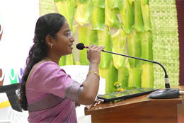 An Indian woman with long hair in a woven silk sari speaks into a microphone with woven wall decoration and large leaves behind her