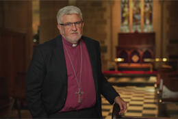 A male bishop with grey beard stands in a stone Cathedral