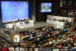 Hundreds of Church of England Synod representatives fill a large hall with a stage and large cross