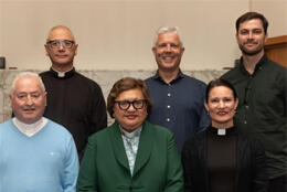 Four clergymmen and two clergywomen stand before a white background