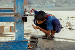 A young boy drinks from a water tank against a sunny dirt road