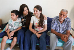 Two children, a woman and a man sit on the edge of a simple bed in an undecorated room