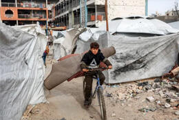 A small boy rides a bike between bombed buildings