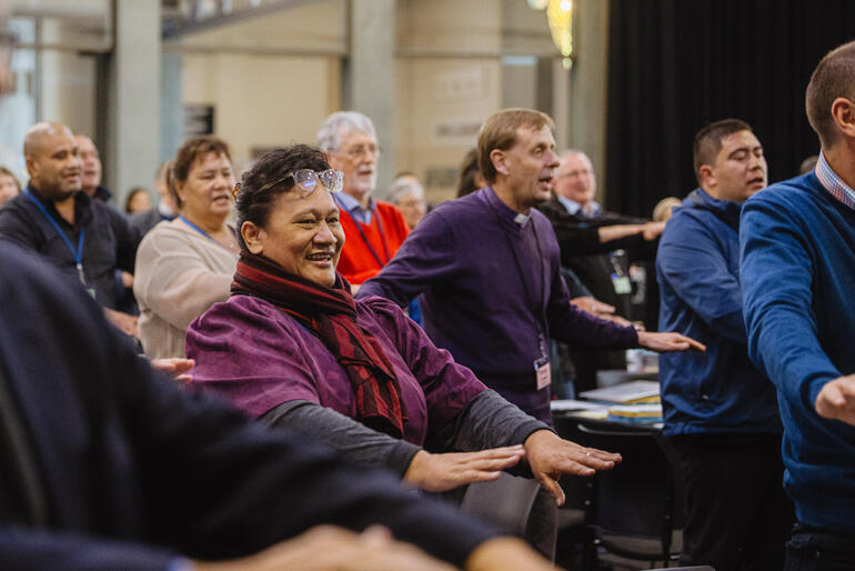 Synod members rise to sing 'Tutira mai ngā iwi' - 'Gather the people together'. Synod members rise to sing 'Tutira mai ngā iwi' - 'Gather the people together'.