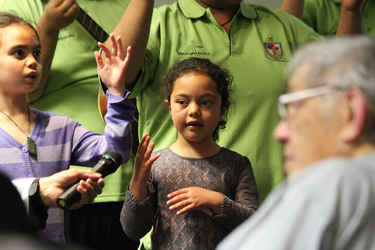 Miss Kaya Walker-Grace Gray goes through the paces of action song. Her koro, Bishop John Gray, is holding the mike.
